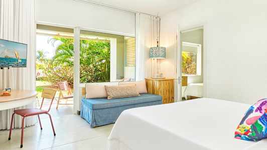 Et lyst hotellrom med seng, sittegruppe og bord. Utsikt mot hagen ses gjennom glassdørene. (AI-generert tekst)