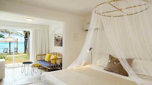 Hotellrom med en stor seng dekket med et myggnett, en sofa, bord og et stort vindu med utsikt over stranden og havet. (AI-generert tekst)