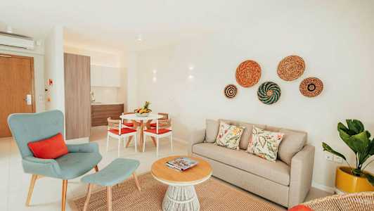 En lys og moderne innredet stue med beige sofa, grønn lenestol og et rundt salongbord av tre. Kjøkkenet er synlig i bakgrunnen. (AI-generert tekst)