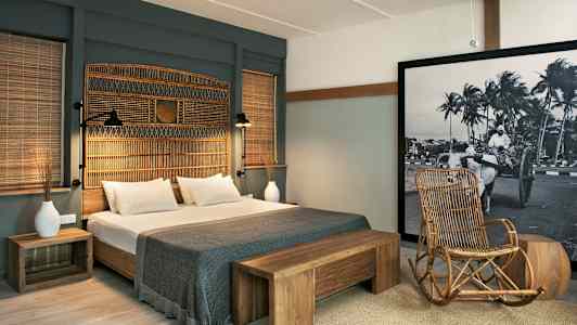 Et hotellrom med en dobbeltseng, tredetaljer og en stol av rotting. Et stort bilde med svart-hvitt motiv pryder veggen. (AI-generert tekst)