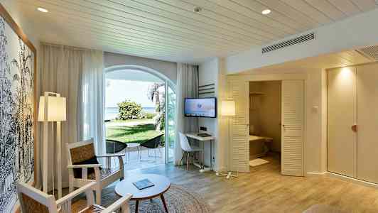 Et hotellrom med sittemøbler, bord og TV. En dør fører til et bad, og det er vinduer som vender mot en grønn hage og stranden. (AI-generert tekst)