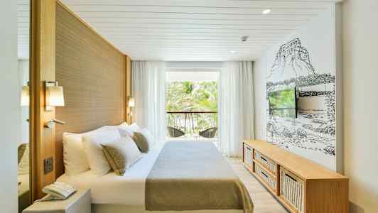 Et hotellrom med en seng, en TV og en balkong med utsikt mot et område med palmetrær. (AI-generert tekst)