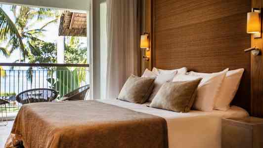 Et rom med en dobbeltseng med beige sengetøy og puter, plassert ved en balkong med utsikt over trær og hav. (AI-generert tekst)