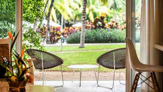 Utsikt fra et rom gjennom en åpen dør mot en uteplass med to stoler og et bord, omgitt av grøntområde og palmer. (AI-generert tekst)
