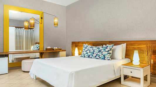 Et hotellrom med en seng med dekorative puter, et nattbord med lampe og et stort speil med gul ramme. (AI-generert tekst)