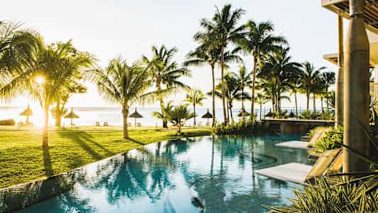 Utsikt over et basseng med solsenger omkranset av palmer og utsikt mot stranden ved sjøen. (AI-generert tekst)