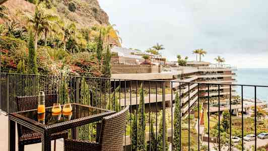 Bilde av en balkong med møbler av rotting, med et bord som har to glass og en karaffel. Utsikt mot planteområder, bygninger og havet. (AI-generert tekst)