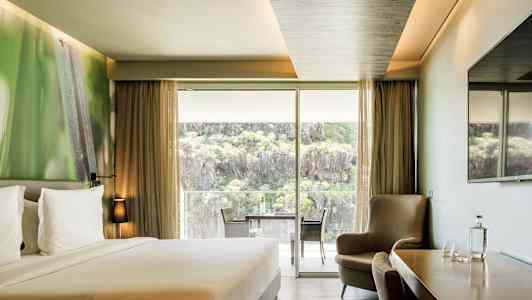 Et hotellrom med en dobbeltseng, sittegruppe, et bord med stoler samt en stor glassdør som åpner opp til en balkong med utsikt over natur og klipper. (AI-generert tekst)