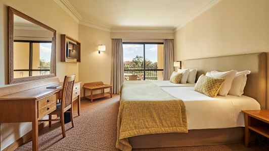 Bilde av et hotellrom med en dobbeltseng, skrivebord og stoler samt en balkong med utsikt. (AI-generert tekst)