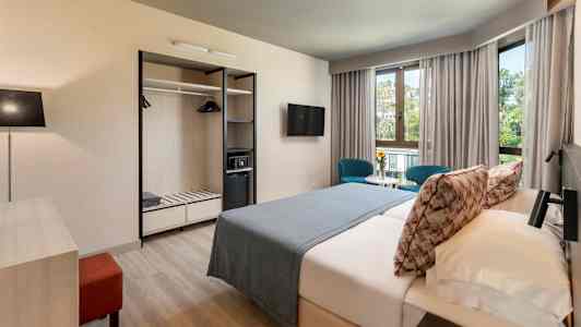 Et hotellrom med dobbeltseng, to blå stoler ved et bord foran vinduer samt garderobe med åpne hyller. (AI-generert tekst)