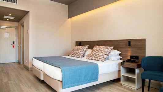 Hotellrom med en dobbeltseng, blå sengeteppe og puter, ved siden av en stol og et nattbord. (AI-generert tekst)