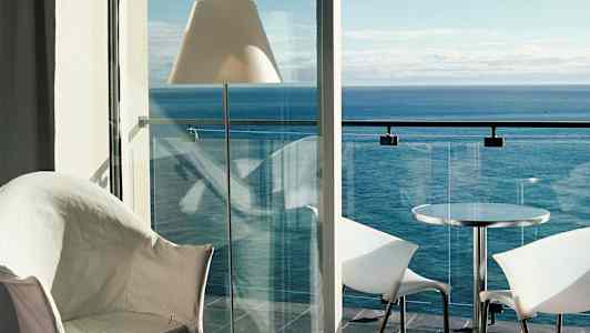 Utsikt fra et rom mot havet, med en balkong med glassrekkverk, bord og stoler. (AI-generert tekst)