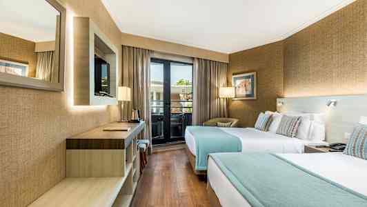 Et hotellrom med to enkeltsenger, en skrivebordsoverflate, en veggmontert TV og et vindu som leder til en balkong. (AI-generert tekst)