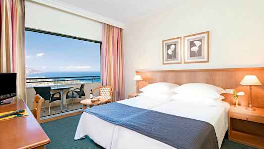 Hotellrom med dobbeltseng, nattbord og balkong med utsikt over havet. (AI-generert tekst)