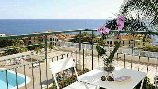 Utsikt fra en balkong med bord og stoler, samt en orkidé i en potte. Et basseng og bygninger i bakgrunnen mot havet. (AI-generert tekst)