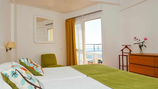 Et hotellrom med to senger, grønne detaljer og utsikt mot en balkong med havet i bakgrunnen. (AI-generert tekst)