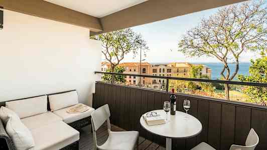 En terrasse med sittegruppe og utsikt mot havet, med bord og to glass vin. (AI-generert tekst)