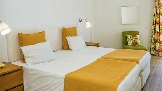 Et hotellrom med to enkeltsenger og gult sengetøy. En grønn lenestol med blomstrete pute og gardiner vises i bakgrunnen. (AI-generert tekst)