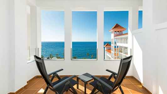 To svarte stoler og et bord på en terrasse med utsikt over havet og en hvit bygning med røde tak. (AI-generert tekst)