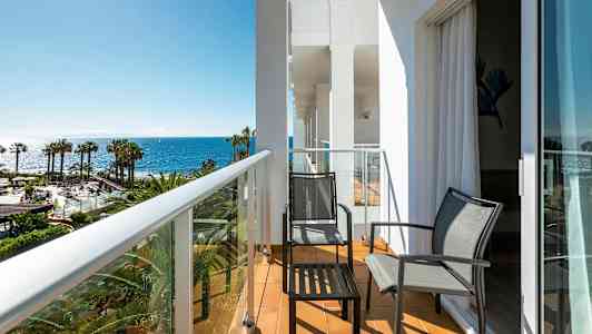 En balkong med to stoler og et bord, med utsikt over palmer og havet. (AI-generert tekst)