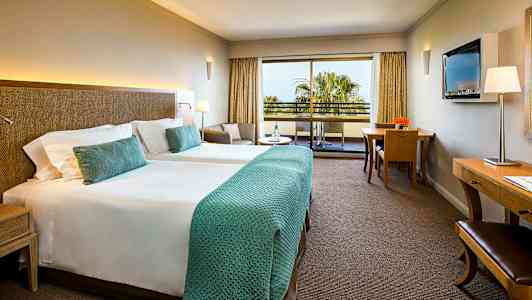 Et hotellrom med to enkeltsenger, en sittegruppe, et skrivebord og en balkong med utsikt. (AI-generert tekst)