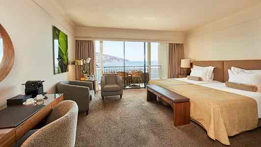 Hotellrom med dobbeltseng, sittegruppe og balkong med utsikt over havet. (AI-generert tekst)