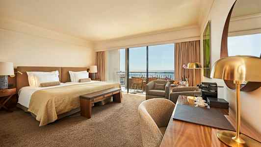 Et stilfullt hotellrom med en dobbeltseng, skrivebord og sittegruppe samt store vinduer mot en balkong. (AI-generert tekst)