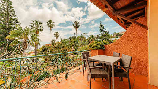 En terrasse med bord og stoler omgitt av grøntområder og palmer. (AI-generert tekst)