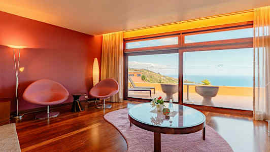 Et rom med røde vegger, to stoler, et bord og store vinduer som vender ut mot en terrasse med havutsikt. (AI-generert tekst)