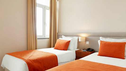 Et hotellrom med to enkeltsenger ved siden av hverandre, med hvite laken og oransje sengetepper og pynteputer. (AI-generert tekst)