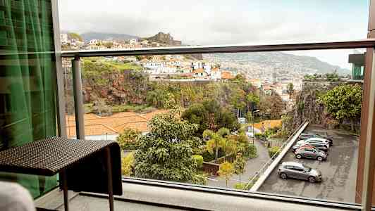 Utsikt fra en balkong med møbler mot et fjellområde med bygninger og parkerte biler. (AI-generert tekst)