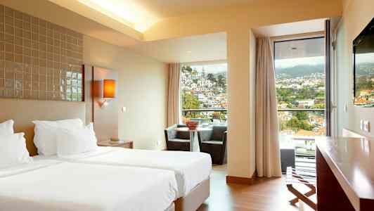 Et hotellrom med to enkeltsenger, en sittegruppe ved et vindu med utsikt over en by i et fjellområde. (AI-generert tekst)