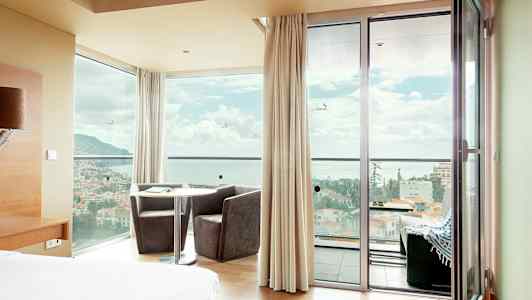 Hotellrom med utsikt gjennom vinduer og dører over en by foran havet. (AI-generert tekst)