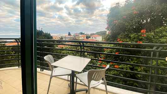 En balkong med et bord og to stoler, utsikt over hustak og trær. (AI-generert tekst)