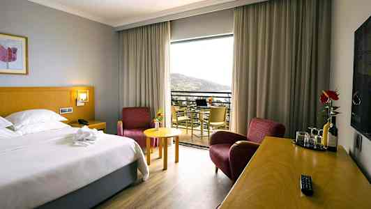 Hotellrom med en dobbeltseng, to røde lenestoler, et skrivebord med drikke og glass, og en balkong med utsikt. (AI-generert tekst)