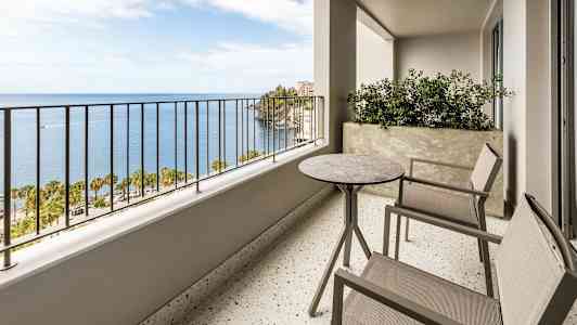 En balkong med to stoler og et bord samt en utsikt over havet og palmer. (AI-generert tekst)