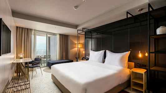 Hotellrom med en dobbeltseng, bord og stoler samt en balkong. (AI-generert tekst)