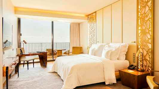 Hotellrom med en dobbeltseng, skrivebord og utsikt over havet gjennom en stor vindusvegg. (AI-generert tekst)