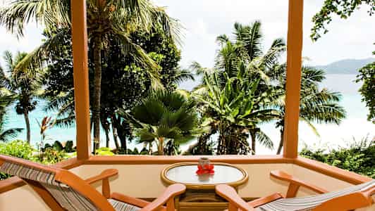 Tremønert terrasse med to stoler og et bord med utsikt over tropiske palmer og blått hav. (AI-generert tekst)