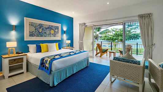 Et hotellrom med en stor seng, blå og gule dekorative puter, en balkong med utsikt over havet og omkringliggende vegetasjon. (AI-generert tekst)