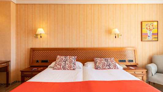 Et hotellrom med en dobbeltseng som har hvitt og oransje sengetøy og dekorative puter. Sengens hodegjerde er i tre med et rutemønstret innfelt. (AI-generert tekst)