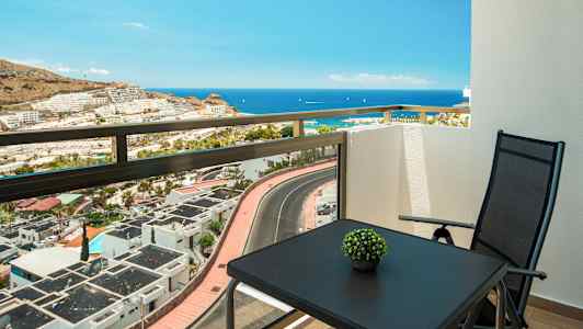 Utsikt fra en balkong mot havet, med et bord og en stol. (AI-generert tekst)