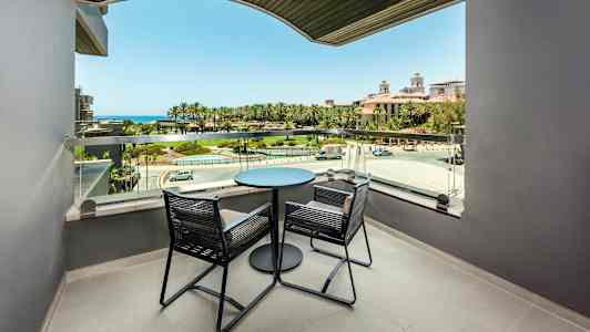 Balkong med to stoler og et bord, utsikt mot grønt område og havet. (AI-generert tekst)