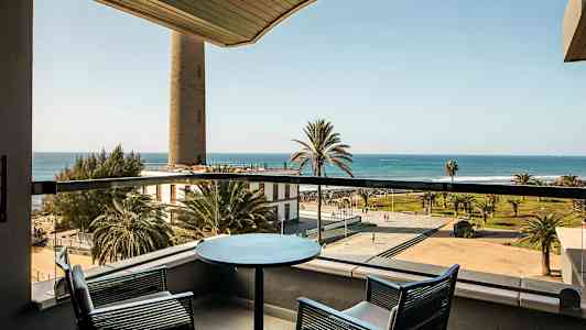 Utsikt fra en balkong med to stoler og et bord mot en strandpromenade, palmer og havet. (AI-generert tekst)