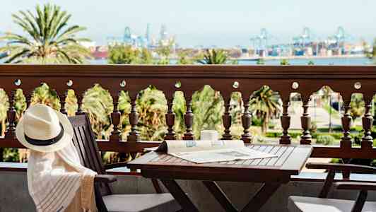 En balkong med bord og stoler, utsikt over trær og industriområde i det fjerne. (AI-generert tekst)