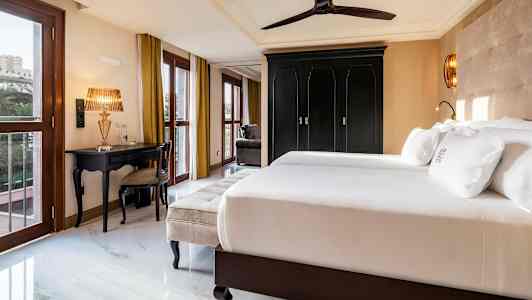 Et hotellrom med dobbeltseng, svart garderobe, gullfarget bordlampe og vinduer med gulldraperier. (AI-generert tekst)