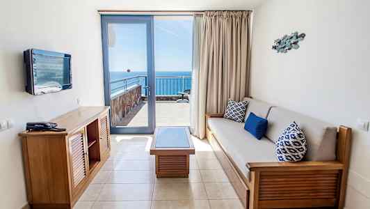 Et hotellrom med sittegruppe og utsikt mot havet fra en balkong. (AI-generert tekst)