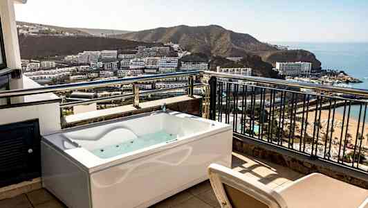 En terrasse med boblebad og utsikt over en bukt med hotell og strand. (AI-generert tekst)