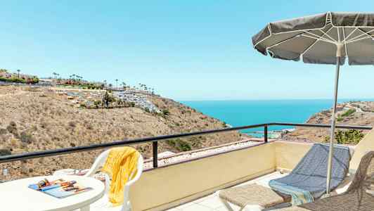 En utsiktsplass med bord og stoler, en parasoll og solsenger som har utsikt over et kystlandskap og havet. (AI-generert tekst)