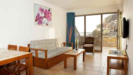 Et stue med treinnredning og utsikt mot en balkong samt bygninger og fjell. (AI-generert tekst)